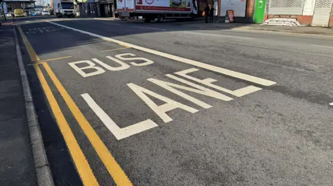 An image of a Bus Lane, with white writing on the floor spelling out 'Bus Lane'. The road is grey and has double yellow lines to the left of the image and is taken on a busy street.