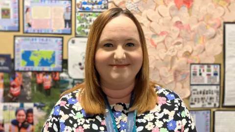 Year 5 teacher Rebecca Laird wearing a floral T-shirt standing in front of a school notice board which is covered in pictures and pieces of work