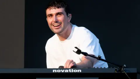 Getty Images Fred Again at Glastonbury, he is wearing a white t-shirt and is leaning over a black DJ deck