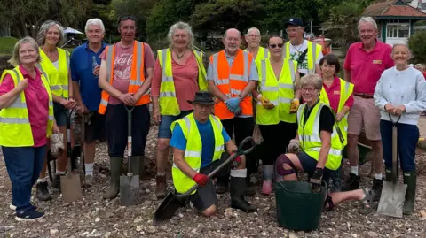 A group of volunteers in high-vis jackets stand and smile with their spades by the brook.
