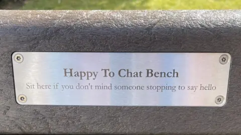 Edd Smith/BBC Plaque on bench which reads Happy to Chat Bench Sit here if you don't mind someone stopping to say hello 