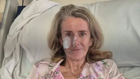 Supplied Laura Corby sitting up in a hospital bed looking directly at the camera. She has a tube up her nose which is taped to her cheek, and is wearing a pale pink floral pyjama top. 