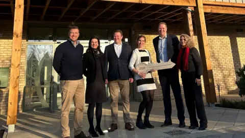 Three men and three women stand in front of a centre. They are all smiling and one is holding a large cardboard key. They are standing in front of a glass-panelled door under an awning. 