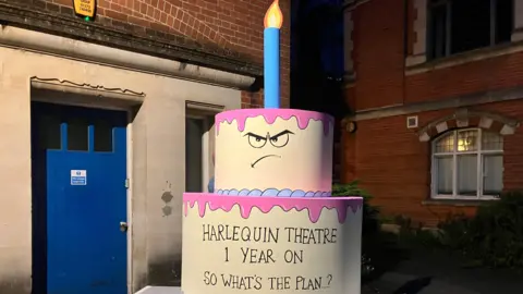 LDRS A model birthday cake with two tiers and a single cake on top. the top tier had an angry face drawn on it while the bottom tier reads Harlequin Theatre one year one So what's the plan?