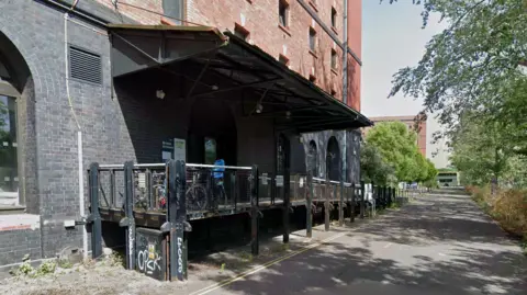 Google A large, black and red brick building with a black metal ramp leading to it. There is a road outside the building flanked by trees. Bikes are chained up outside the building. 