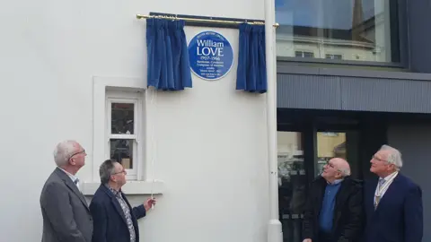 A group of people stand around the blue plaque dedicated to Love. The blue circular plaque is high up on a white wall and is surrounded by blue curtains which have just been drawn back. 
