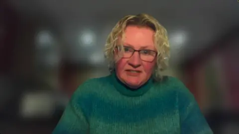 Catherine Clarke has a green jumper, blonde curly hair and glasses.