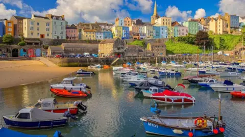 A wide shot of Tenby harbour, with colourful buildings nestled in the cliff overlooking the water. A number of boats are lined up at the front of the shot with the beach on the left hand side.