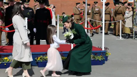 Reuters A woman wearing a bottle green coat with an Irish Guards cap star pinned to it crouching down to greet a young girl wearing white at a St Patrick's Day parade