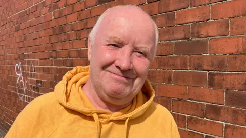 Jennie Dennett Stuart Benson, a balding middle-aged man with short, white hair. He is wearing an orange hoodie and stands next to a brick wall.