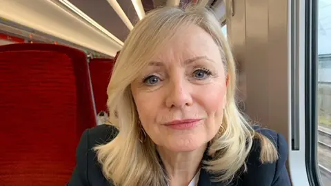 WYCA Tracy Brabin looks into the camera. She has long blonde hair and is wearing a navy jacket. She is sitting on a train seat.