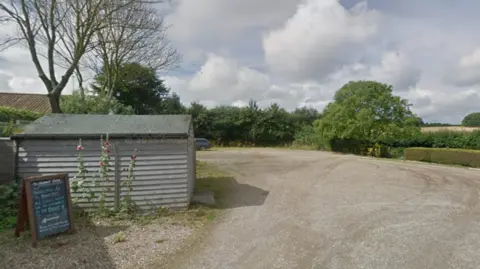 Google A gravel car park lined with hedgerow and trees, with a shed in the foreground on a bright cloudy day.