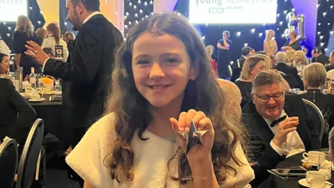 Articulate PR A young girl with brown wavy long hair at an award ceremony holding an award, with a gold star on it. She is surrounded by tables and chairs with people in formal clothes.