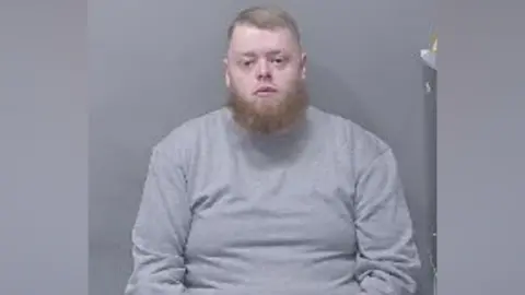 A custody image of Tyler McQuarrie, wearing a grey top and sitting looking straight at the camera. He has a ginger coloured beard and short fair hair.
