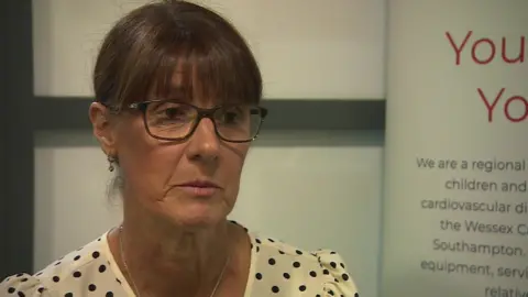 Sharon Brady wearing spotted dress and glasses, sitting in front of white background.