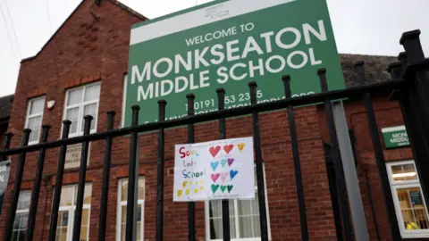 LDRS An exterior view of a red brick school with black railings outside. A large green sign with white text reads 'Monkseaton Middle School' and a hand-drawn picture attached to the railings reads 'Save this school'.
