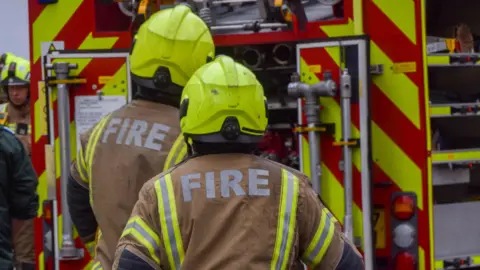 Two firefighters wearing brown uniforms that say in white letters 'Fire' on the back. They are wearing high vis hard hats and are stood near a fire engine. 