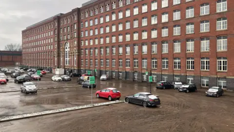 Meadow Mill in Stockport seen from the street, with mud covering the car park, where there are few snow-covered car. 