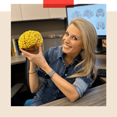 Lara Lewington sitting at table holding a 3D printed gold coloured model of her own brain