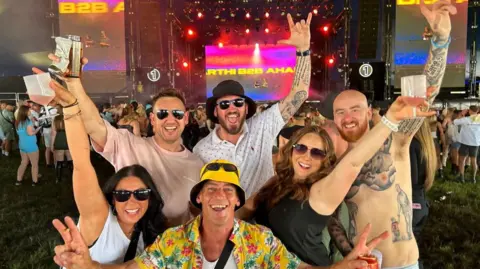 NNP Six people gathered together and smiling into the camera with their arms in the air. They are standing in front of one of the stages at the festival. There are four men and three women and most of them are holding a drink in their hands. One man is crouched down in the front of the group and is wearing a yellow bucket hat and yellow Hawaiian shirt and is holding both of his hands up in peace signs. The two women stand either side of him with dark hair and sunglasses on and the three men stand behind them. The man on the right is shirtless with tattoos across his torso. 