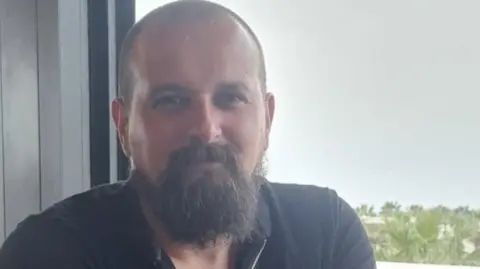 A bald man with a beard and dark shirt smiles as he stands in front of a window.