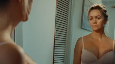 Breast Cancer Now A woman looking at herself in the mirror. She is wearing a white bra.