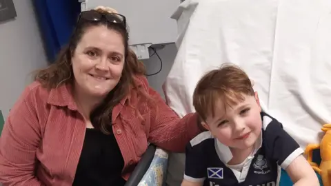 Rebekah, a woman with brown hair, wearing a pink shirt, sat with her young son Hector who looks up at the camera in a hospital room.