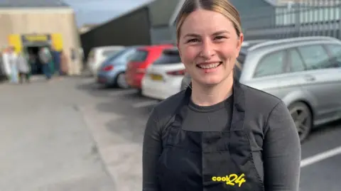 Ffion Roberts, wearing an apron with Cook24 written on it, smiles at the camera outside the site of the cooking course 