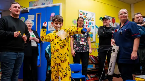 Warrington and Halton Teaching Hospitals Charity A child in a bright yellow Pikachu onesie stands in a hospital playroom, arms raised throwing a magnetic dart. Luke Littler is behind him holding several more magnetic darts. Several adults and staff members stand nearby, some holding small toys or medical equipment. The walls are decorated with posters and learning materials.