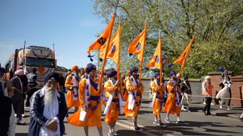 Sikhs wearing orange and purple outfits carry flags of the same colour during a street parade, with a lorry behind them decked out with the colours as part of the procession.