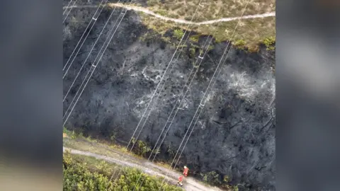 Surrey Fire and Rescue Service An aerial view showing two people walking along a path next to a large area of burnt green land. Both are wearing high viz orange outfits.