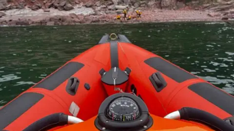 The bow of an orange RIB at sea with a number of people on a beach.