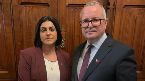 Lee Barron Home Secretary Shabana Mahmood standing next to Corby and east Northamptonshire MP Lee Barron. Mahmood is wearing a white blouse with a burgundy suit jacket over the top. She is looking directly at the camera. Barron is wearing a suit jacket with a white shirt and purple tie. He is wearing glasses and is looking directly at the camera.