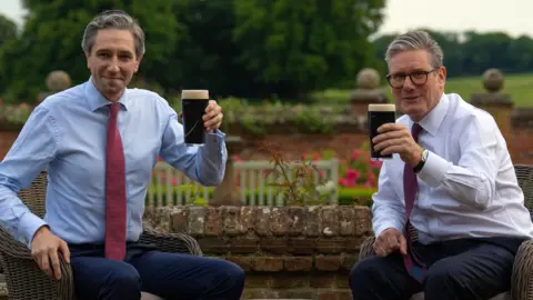 PA Media Sir Keir Starmer (right) and Simon Harris drink a pint of Guinness during his visit to Chequers. Starmer has grey hair, wearing glasses, a white shirt burgundy tie, and dark trousers. Harris has grey hair, wearing a light blue shirt, burgundy tie and navy trousers. Both are holding up a pint of Guinness.