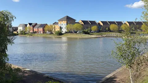 Ben Schofield/BBC A shot across a man-made lake towards a collection of fairly new housing. The image is framed by small trees and is shot from the corner of the lake. On the other side of the water, a grassy bank reaches out of the water, behind which are houses, bathed in bright sunlight. One of the buildings is four storeys high, while others are three and two-storey structures. The sky is blue, save for two small puffy clouds.