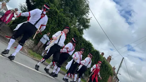 A group of men and women in traditional clothing grasp wooden poles as they dance in a residential street.