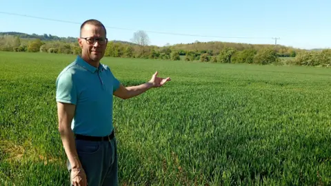 Stewart Long with short dark hair, looking at the camera and wearing a light blue T-shirt and brown trousers. He is pointing with his left hand across a field with tall grass and a hedge in the background.