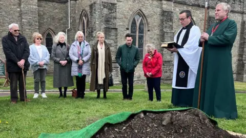 A group of people stand on grass outside a stone church building, gathered around a small mound of earth covered with green fabric. Two people wearing clerical robes stand at the front, one holding an open book, while others stand nearby with heads lowered. 