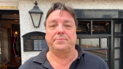 A man with short, brown hair wearing a black polo shirt. He is standing in front of a pub.