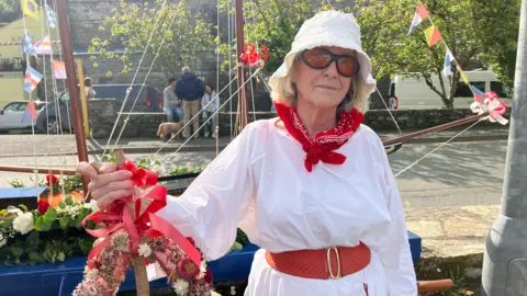 A woman wearing a white dress and red neck scarf is standing next to a wooden boat structure. She is wearing a hat and glasses and is looking toward the camera. A road with trees and a stone wall are in the background behind her. 