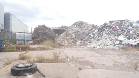 EA Two large piles of rubbish and/or waste can be seen with no cover on. A tyre is left on the ground in the foreground. An old tree is next to one pile and weeds grow out of the ground nearby.