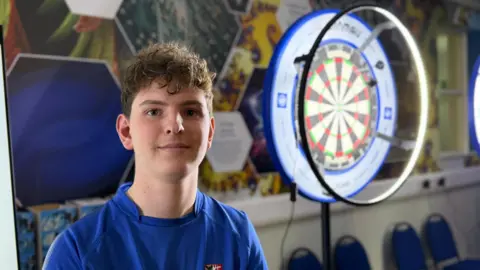 Elliot Deady/BBC A boy in a blue top on with a brightly-lit dartboard in the background