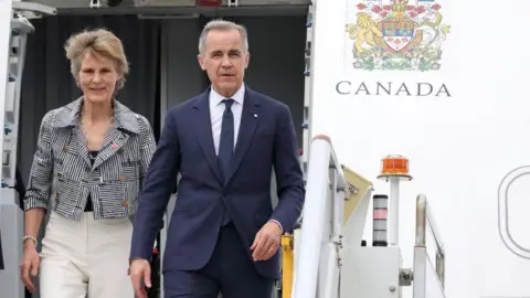 Mark Carney and his wife, Diana Fox Carney, at the top of the airplane stairs. A Canada logo is to their right. 