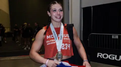 @inov8 A woman with dark hair tied back in plaits, with a large gold medal around her neck, over a red top, and holding a Union Flag, smiles as she walks through a large hall.