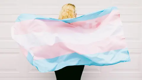 A transgendered person with long blonde hair faces away from the camera and holds the blue, pink and white trans flag behind their back. 