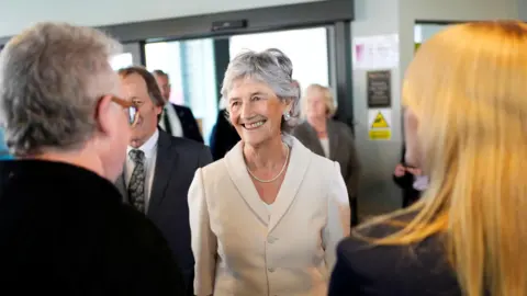 Catherine Connolly can be seen smiling, engaging with people. She is wearing a white suit jacket and a white pearl necklace as well as silver earrings.