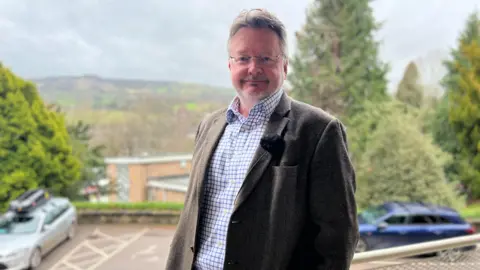 Man in cheque short and brown tweed jacket stands on balcony infront of car park and hillside