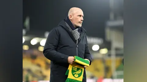 Norwich City Philippe Clement is wearing a scarf and black padded coat. He is standing on the pitch and has a Norwich City scarf in his hands.