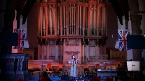 Paul Rodgers Maz O'Connor is playing a guitar in front of a large organ with two Union Jack flags hanging to either side of it. The organ and O'Connor are being gently lit.