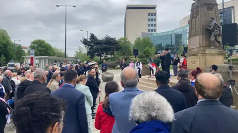 A crowd of men and women in uniforms and civilian dress looking towards the stone Bradford War Memorial.